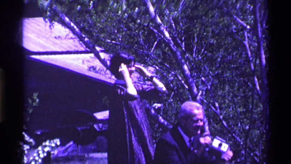 A man in a dark jacket holds a camera outdoors in the Ohio home-movie scene, with trees and a house visible behind him.