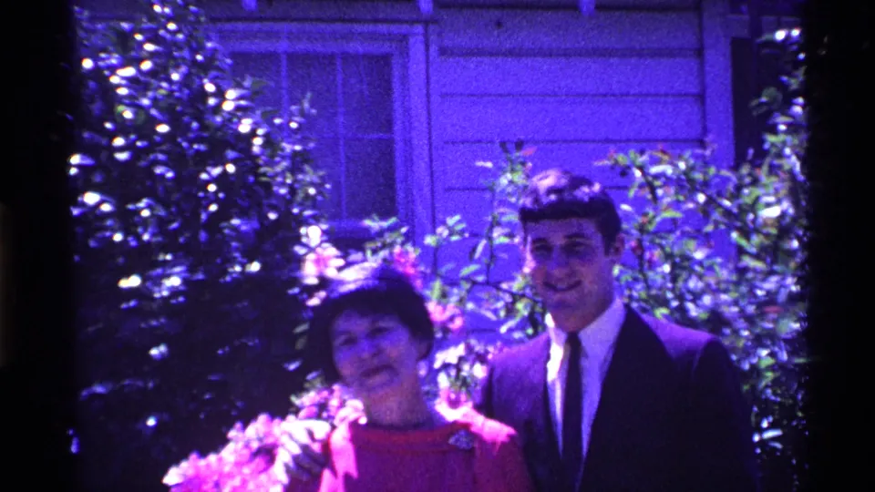 The woman in a red dress and the young man in a dark suit smile together in an Ohio backyard garden in this 1969 home movie.