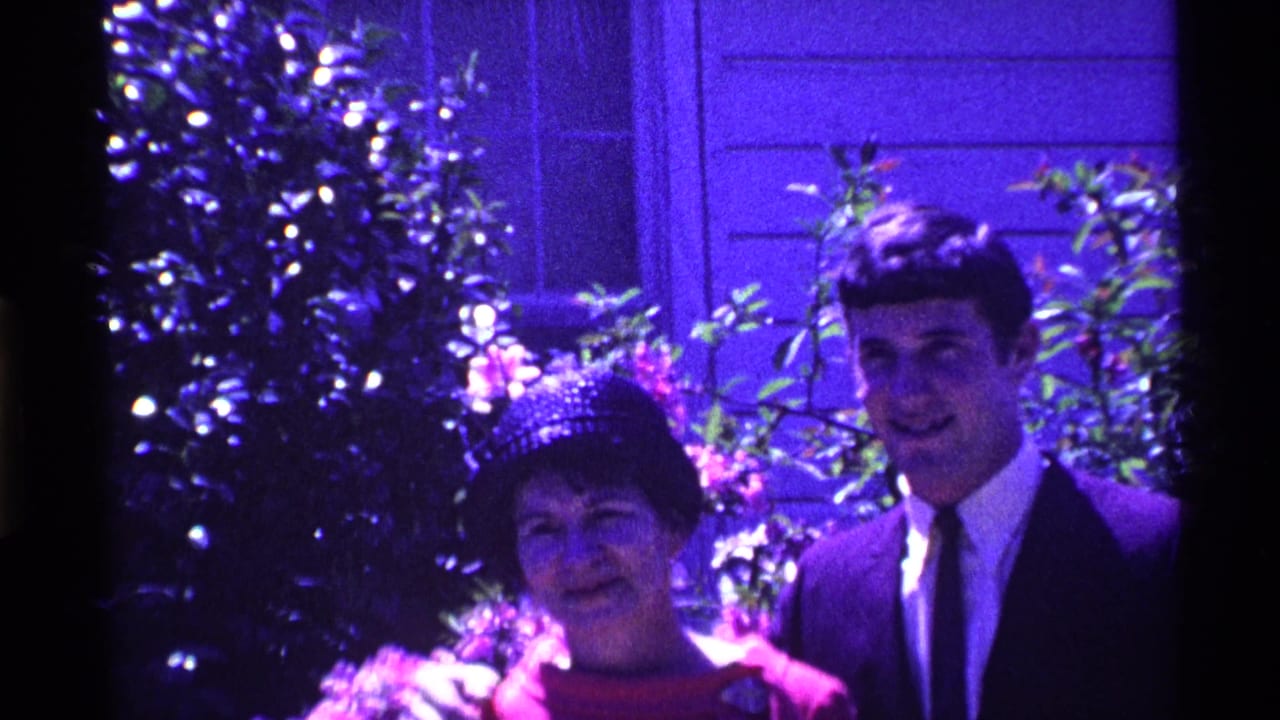 Woman in red dress and hat standing beside young man in dark suit outdoors.