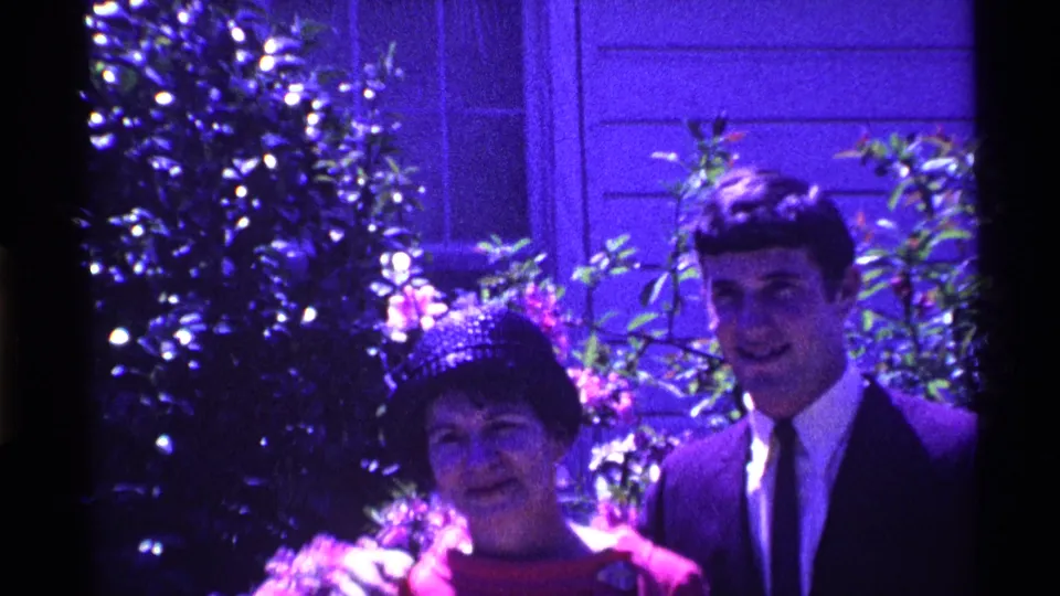 In 1969 Ohio home-movie footage, a woman in a red dress and patterned hat stands beside a young man in a dark suit near shrubs and a house.