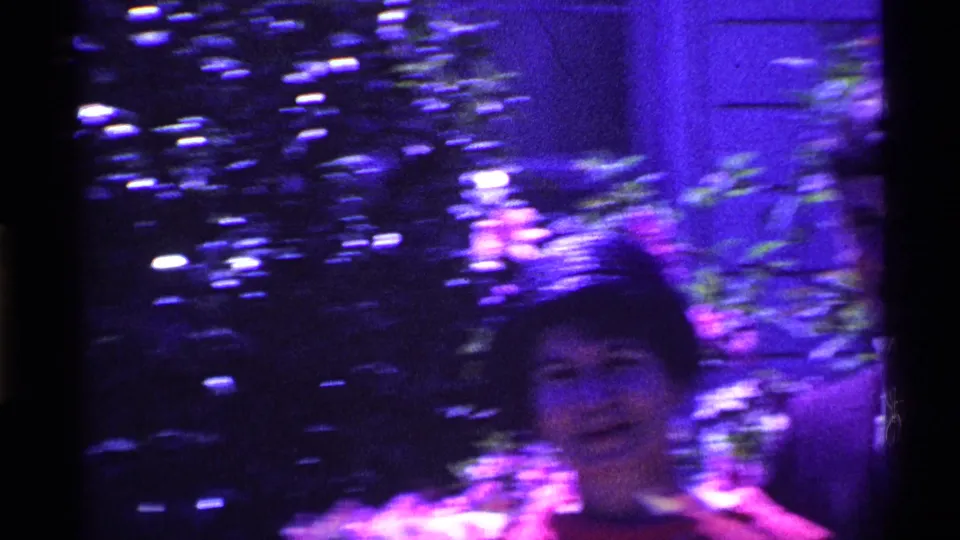 A motion-blurred frame from the 1969 Ohio 8mm clip shows the woman in red near leafy shrubs, with the young man mostly out of view.
