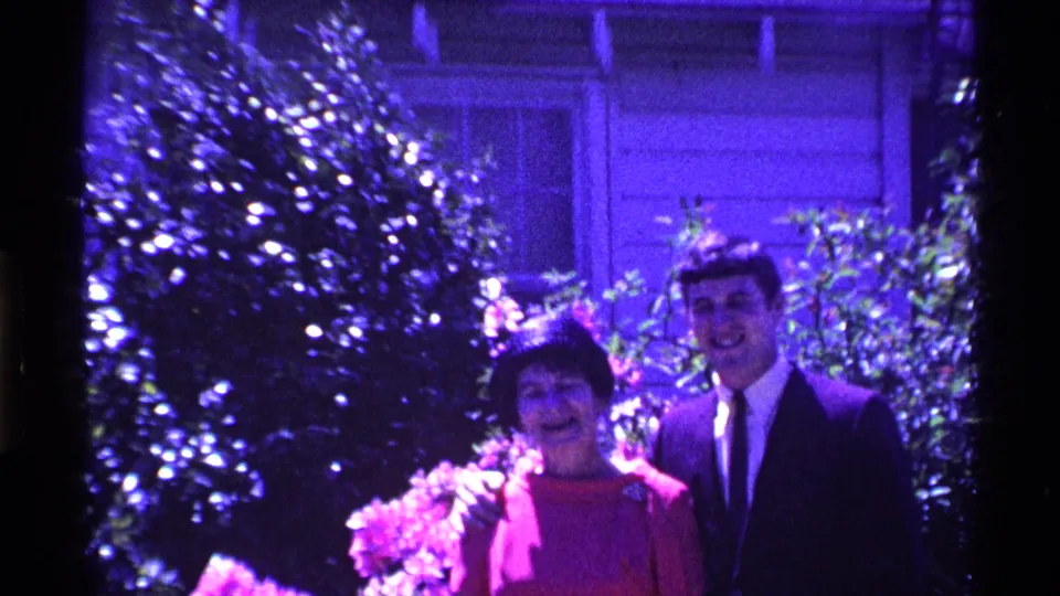 The woman and young man smile toward the camera in an Ohio 1969 home-movie frame, surrounded by bright foliage and a house wall.
