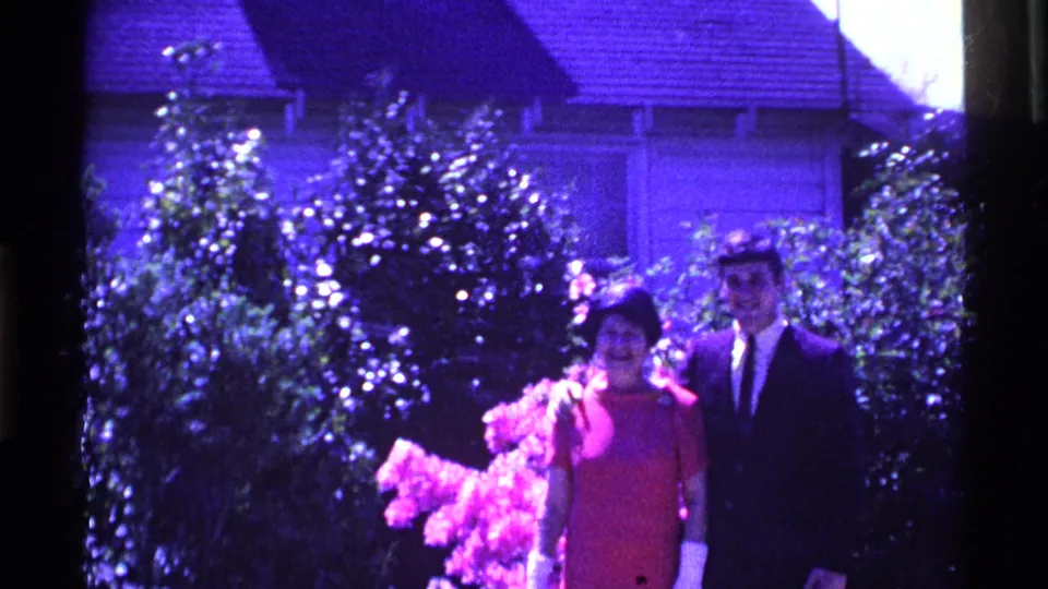 A wider 1969 Ohio home-movie view shows the woman in red and the suited young man standing beside flowering shrubs outside a house.