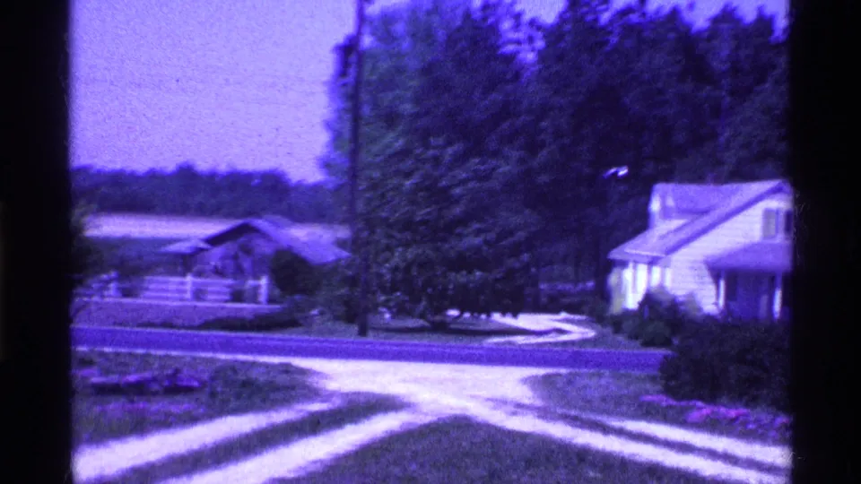 1969 Ohio 8mm view of a curving driveway, white house, trees, white fence, open field, and low structure beyond.