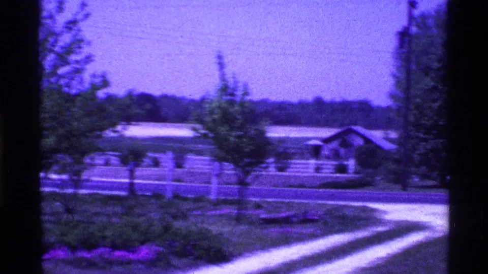 1969 Ohio 8mm frame showing a white fence along an open field, with trees, a driveway edge, and a low outbuilding in the background.