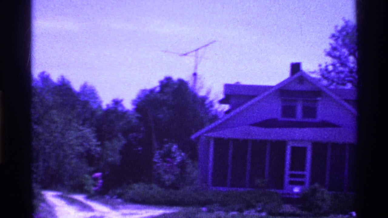 Purple-tinted 8mm frame of a small house with a screened porch beside a dirt driveway.