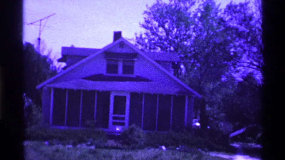 1969 Ohio home-movie frame centered on a modest house with screened porch, chimney, rooftop antenna, lawn, and surrounding trees.