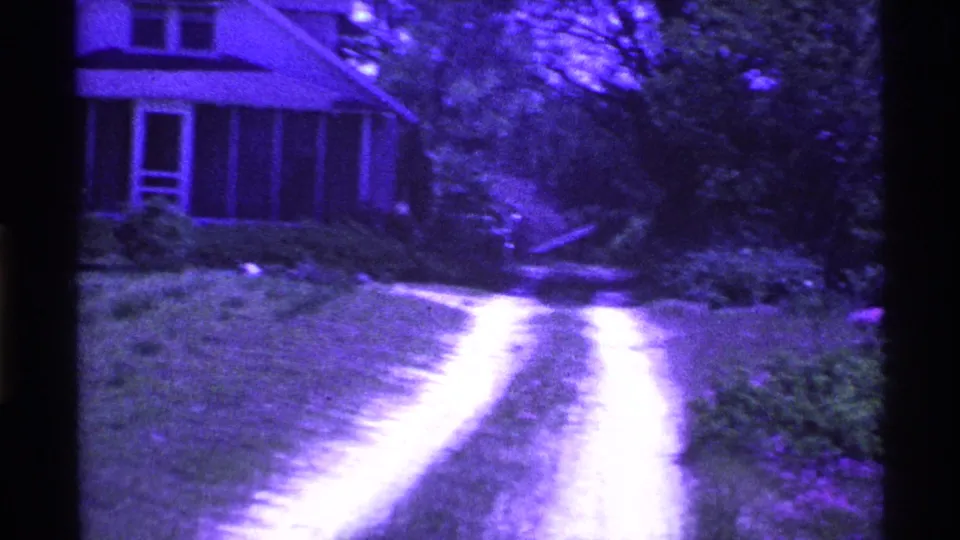 1969 Ohio home-movie frame with a small house, grassy yard, bright dirt driveway, and surrounding trees under a purple-shifted film image.