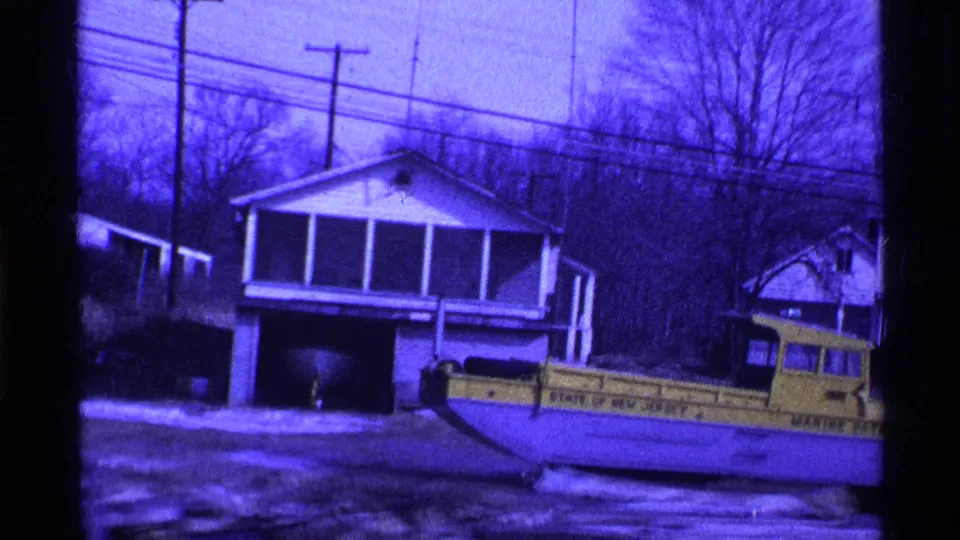 1969 Ohio home-movie frame showing a snowy yard, porch-front houses, utility poles, and a yellow state marine patrol vehicle.