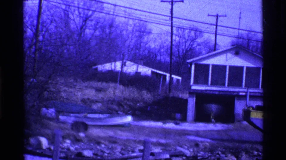 1969 Ohio frame showing a snowy residential yard, a small boat-like craft, houses, bare trees, utility poles, and part of a yellow patrol vehicle.