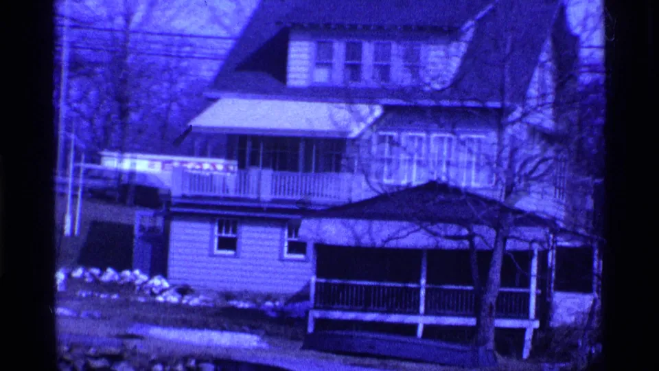 1969 Ohio 8mm frame showing the side and porch of a suburban house, with snow patches and leafless branches across the foreground.