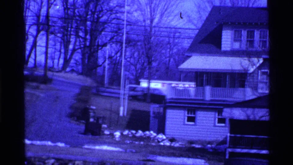 1969 Ohio 8mm frame showing a house at right, sloped driveway at left, bare trees, utility pole, snow patches, and dark bins near the yard.