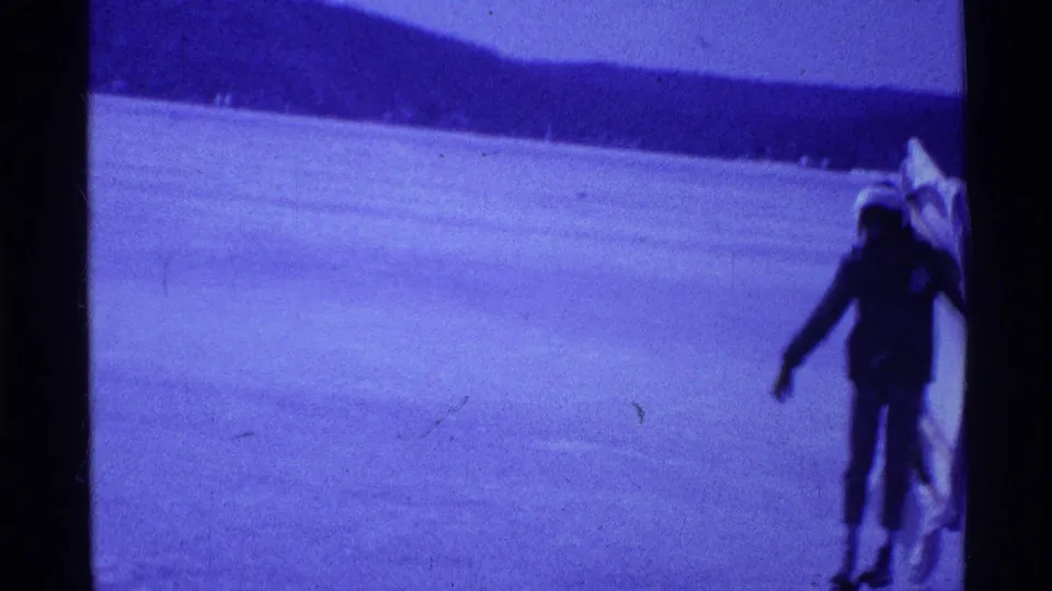 1969 Ohio frame showing a distant person in dark winter clothing carrying a sail or board-like object across a frozen lake.