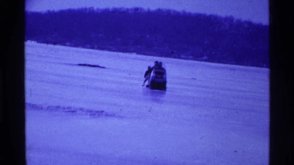 1969 Ohio home-movie frame of a snowmobile and riders crossing a frozen river or lake bed toward the camera.
