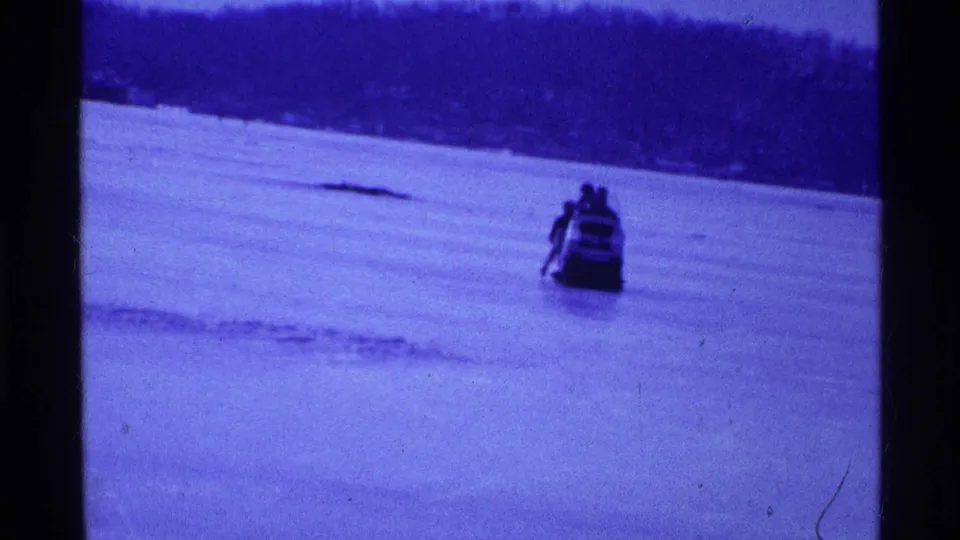 Blue-tinted 1969 Ohio 8mm frame with a snowmobile moving over a frozen river or lake bed near a wooded shore.