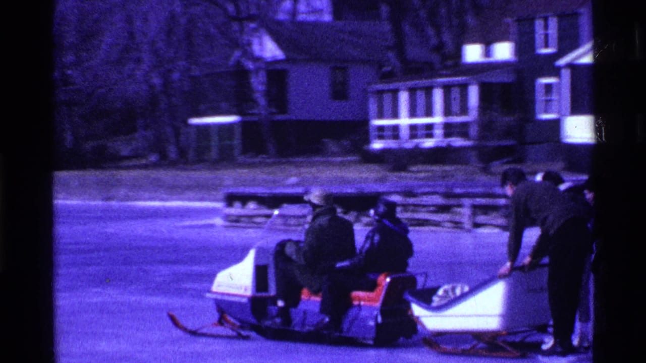 In 1969 Ohio home-movie footage, bundled people stand and sit by a snowmobile and sleds on a frozen lake with houses beyond.