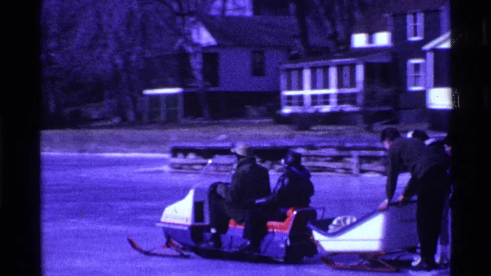 In 1969 Ohio home-movie footage, bundled people stand and sit by a snowmobile and sleds on a frozen lake with houses beyond.