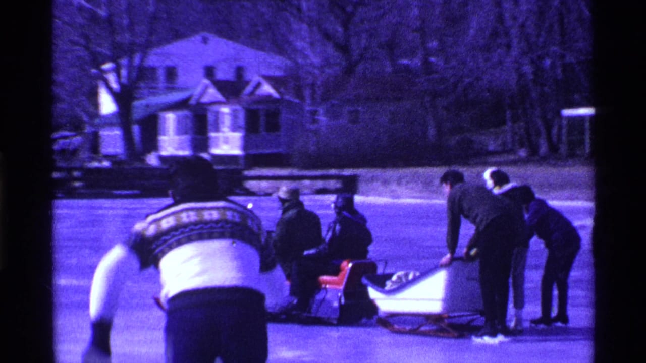 Bundled people on an Ohio frozen lake gather around sleds and a snowmobile, with one figure bending forward in the foreground.