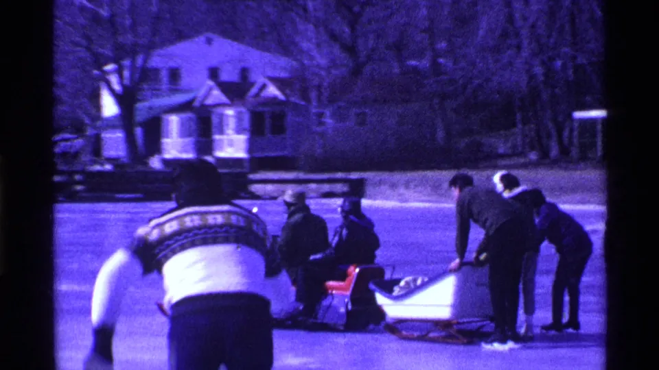 Bundled people on an Ohio frozen lake gather around sleds and a snowmobile, with one figure bending forward in the foreground.