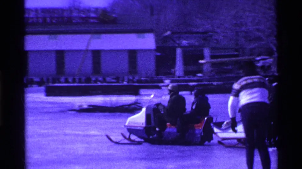 A group in winter clothing sits on a sled on the frozen lake, with a standing figure nearby and a boathouse-like building behind.