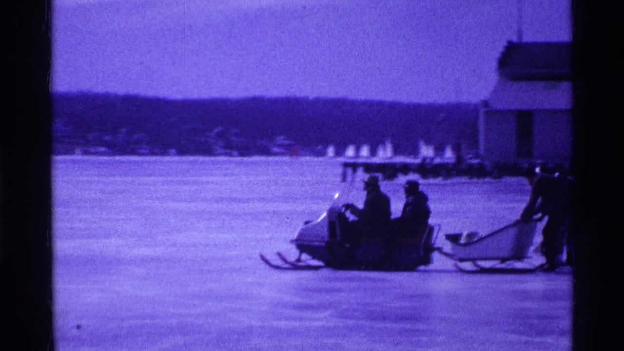 Snowmobile with riders and a tow sled on a frozen lake near a distant shoreline.