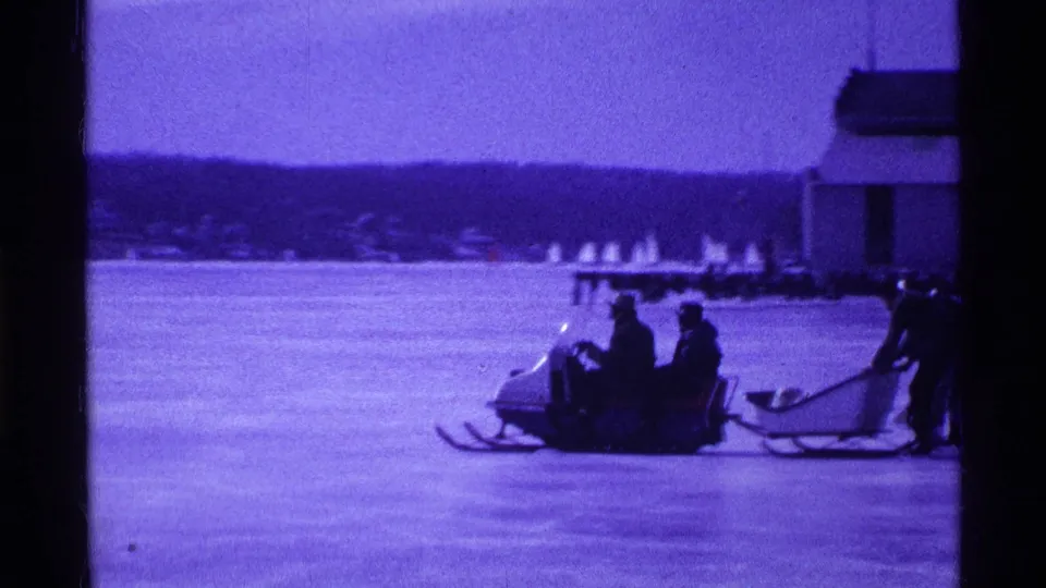 1969 Ohio home-movie frame showing bundled people on a snowmobile and sled crossing a frozen lake.