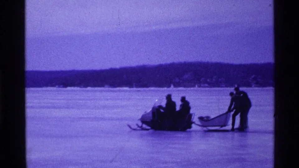 Distant 1969 Ohio winter recreation scene with people riding a snowmobile and sled on a frozen lake.