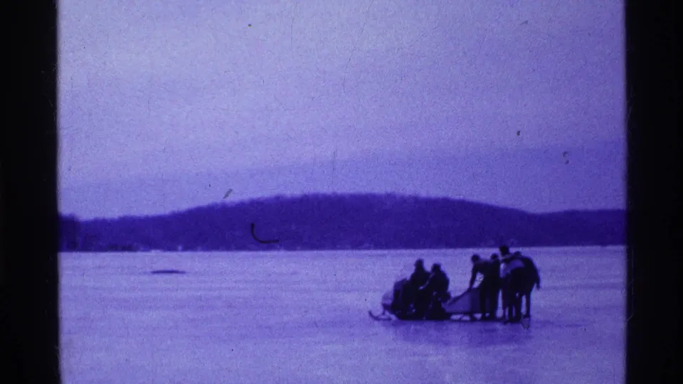 Wide 1969 Ohio home-movie view of a snowmobile outing on a frozen lake with wooded hills behind.