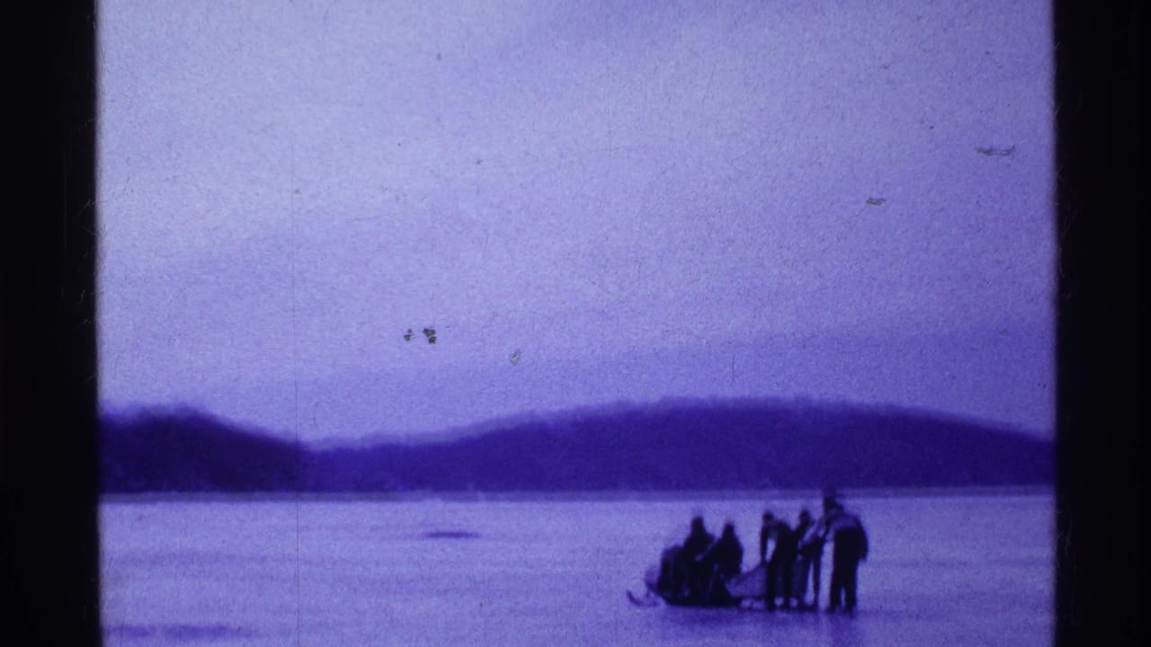 1969 Ohio home-movie frame showing people gathered around a sled on a frozen lake with wooded hills in the distance.