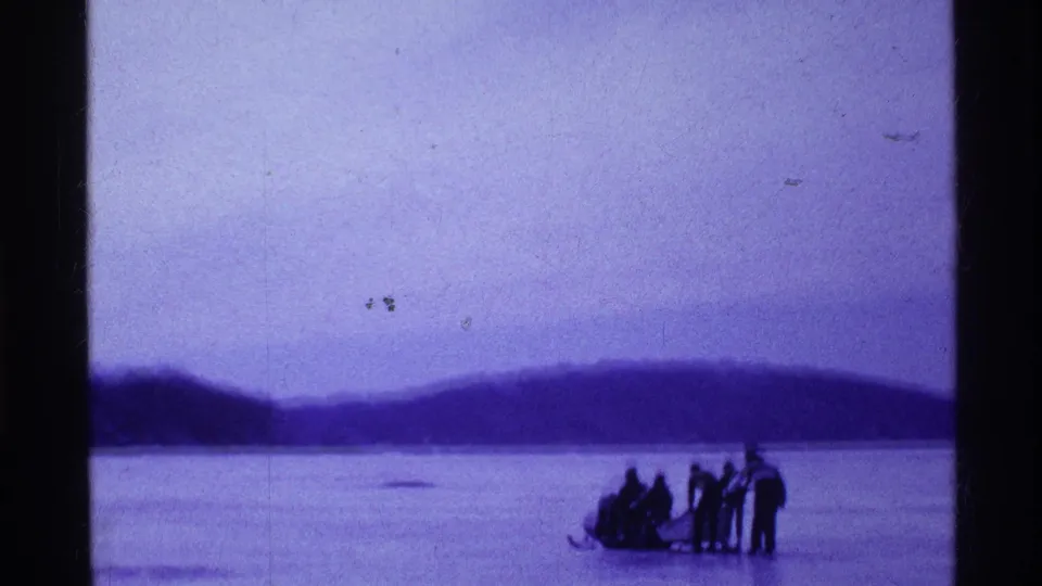 1969 Ohio home-movie frame showing people gathered around a sled on a frozen lake with wooded hills in the distance.