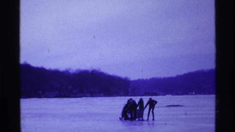 Distant 1969 Ohio home-movie view of people beside a sled on frozen lake ice, with low hills and shoreline behind them.