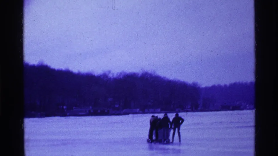 1969 Ohio winter frame showing a small group and sled on frozen lake ice, filmed with dark tree-lined hills behind them.
