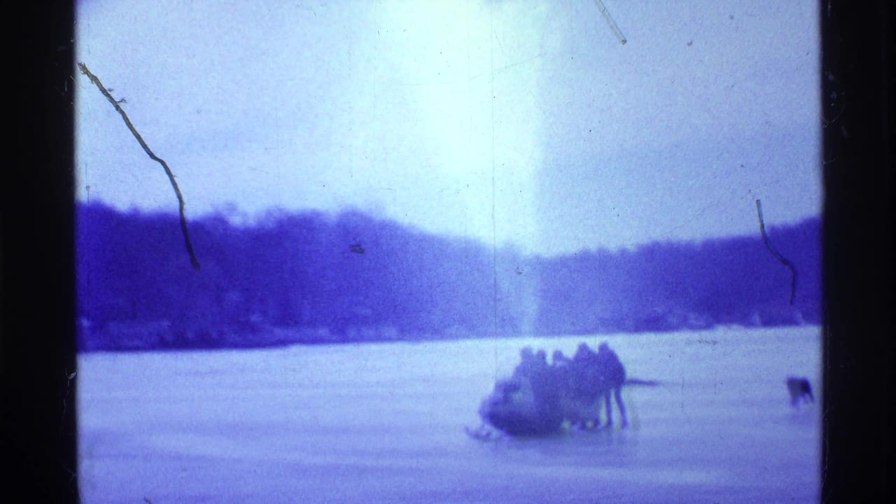 Distant group with a snowmobile on a frozen lake near a wooded shore.