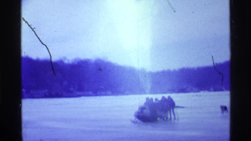 1969 Ohio home-movie frame showing people gathered around a snowmobile on frozen lake ice with a dark tree-lined shore behind them.