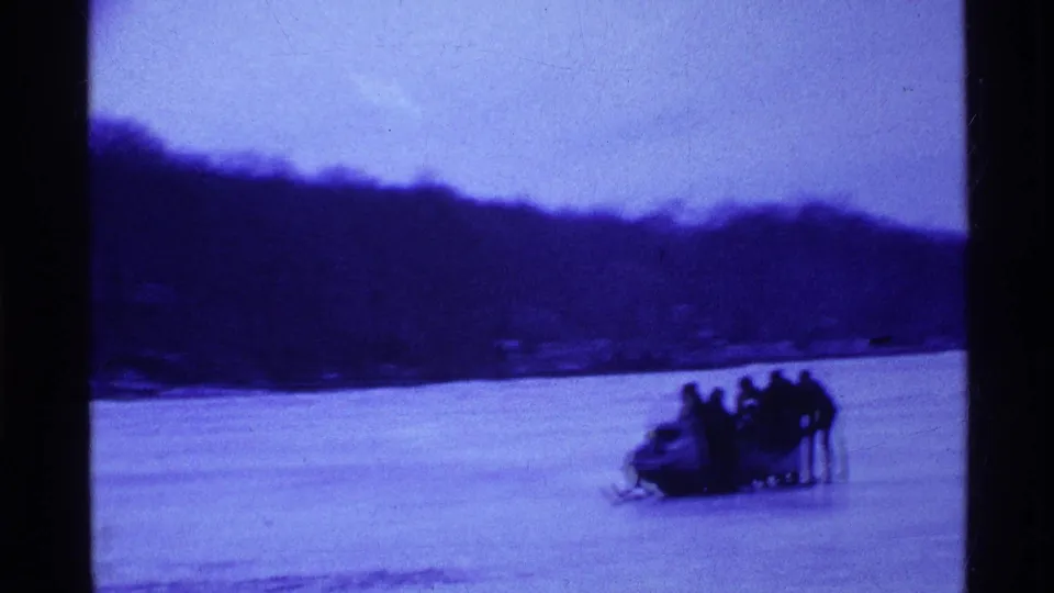 Blue-tinted 8mm frame of a snowmobile with several people on or beside it moving across an Ohio frozen lake in 1969.