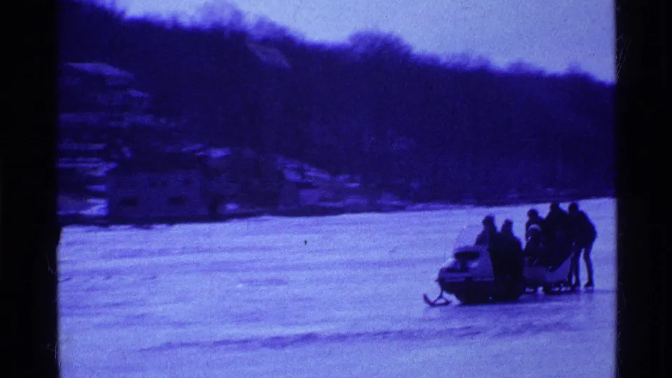 1969 Ohio winter outing frame with a group clustered near a snowmobile on frozen lake ice below a dark wooded shoreline.