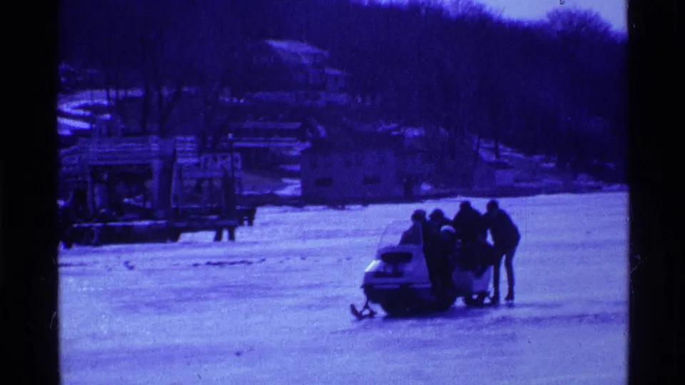 Blue-shifted 8mm frame from 1969 Ohio showing people gathered by a snowmobile on ice with lakeside structures and trees in the background.