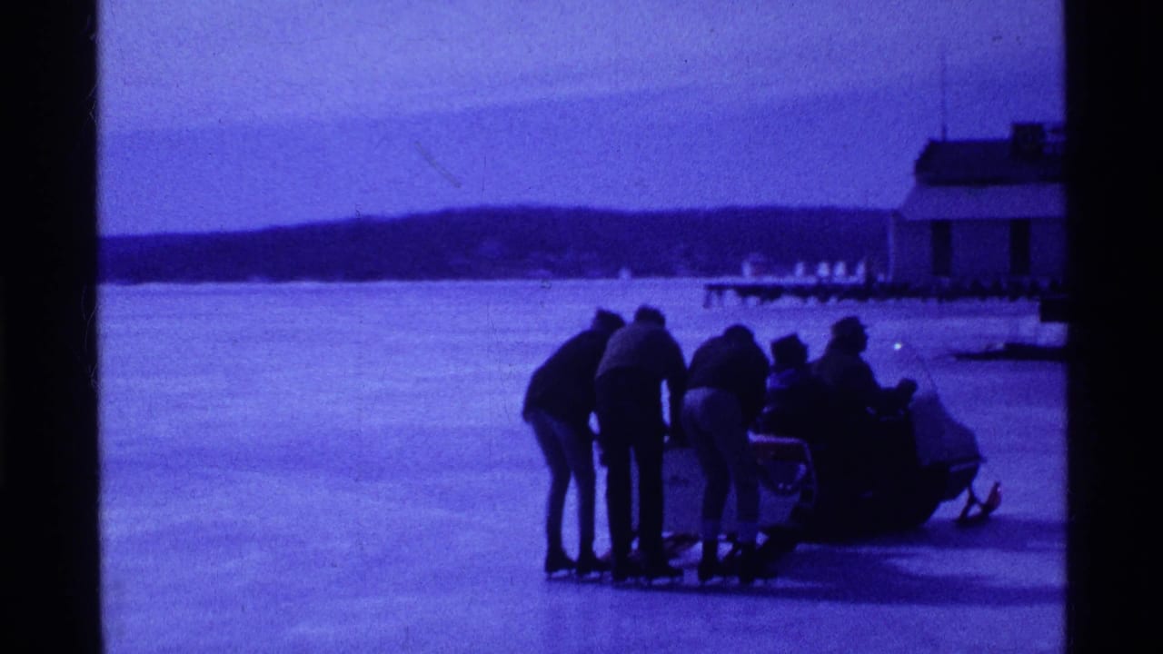 1969 Ohio home-movie frame showing bundled people on a frozen lake gathered around a small sled or snow vehicle.