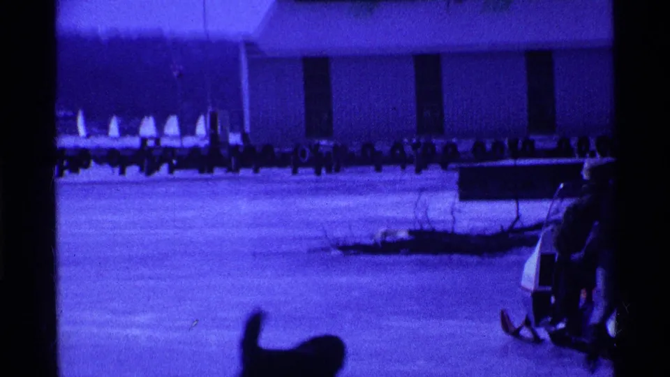 A dog moves through the foreground while bundled people and winter equipment are partly visible near the frozen lake edge.