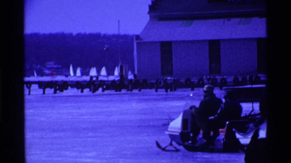 Two bundled people sit on a sled or snow vehicle on the frozen Ohio lake, with docked sailboats and a building behind them.