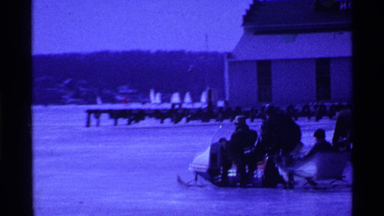 In 1969 Ohio 8mm footage, people in winter clothing gather around a sled on a frozen lake beside a building and distant shoreline.