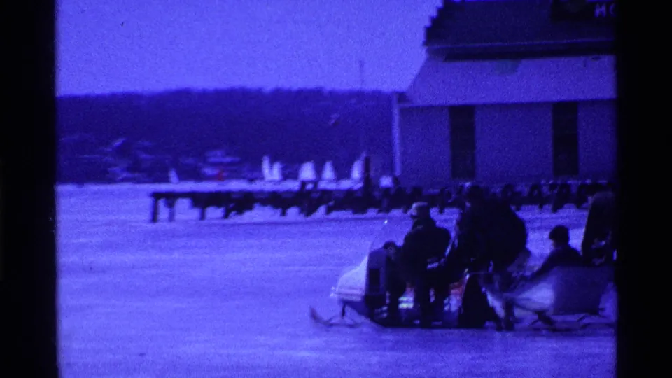 In 1969 Ohio 8mm footage, people in winter clothing gather around a sled on a frozen lake beside a building and distant shoreline.