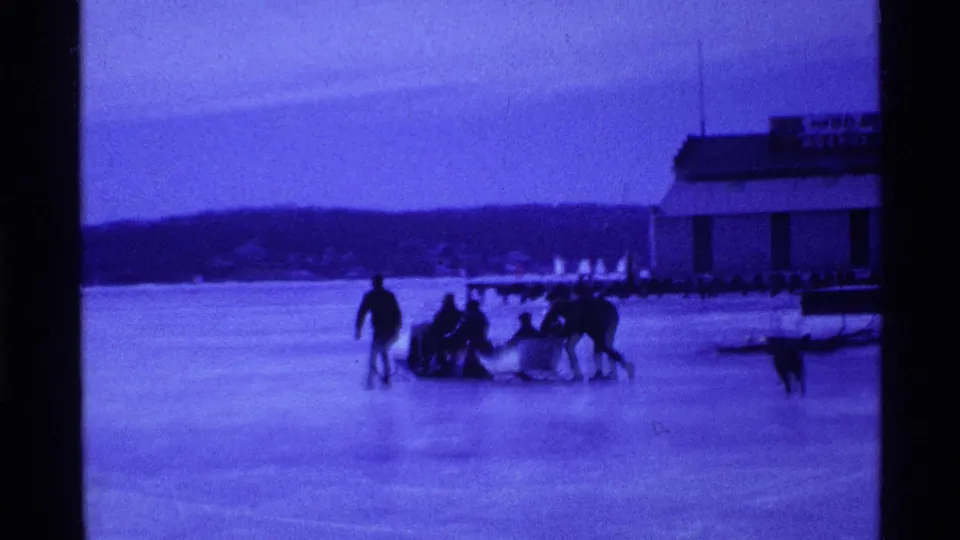 People move a sled across the frozen lake in Ohio as a dog runs near the group, with shoreline and a signed building in the background.