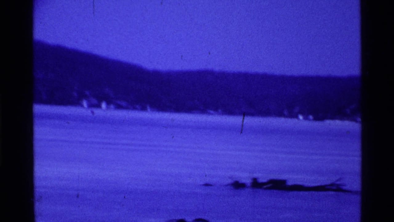 Blue-tinted 1969 Ohio 8mm frame showing a frozen lake, wooded hillside, and distant people on the ice.