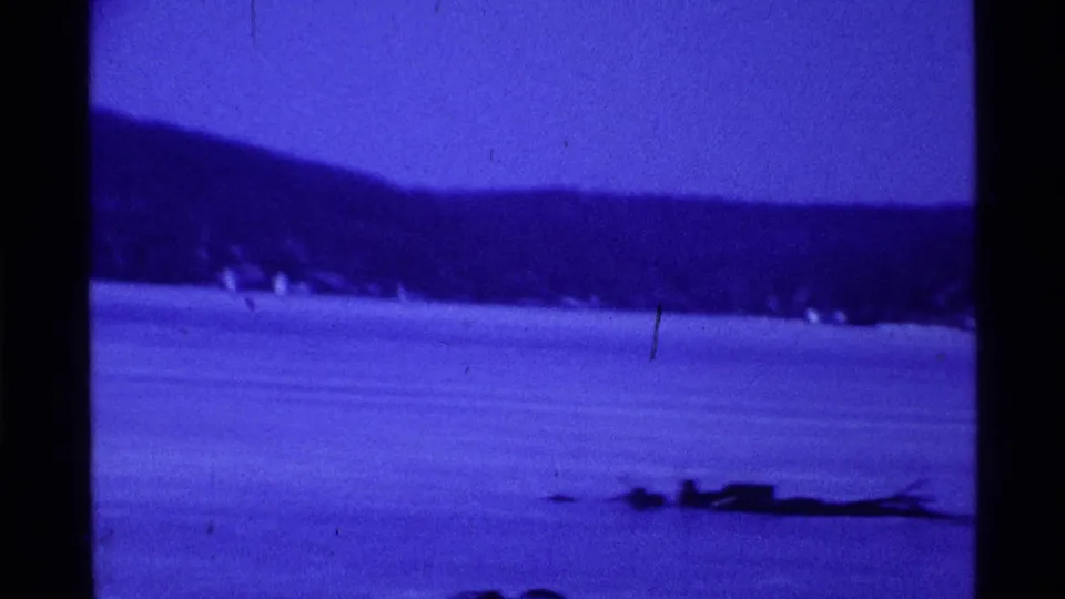 Blue-tinted 1969 Ohio 8mm frame showing a frozen lake, wooded hillside, and distant people on the ice.