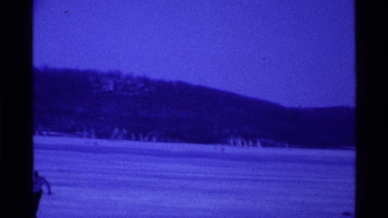 Blue-cast 8mm view of an Ohio frozen lake in 1969, with distant figures and a dark tree-lined hill.