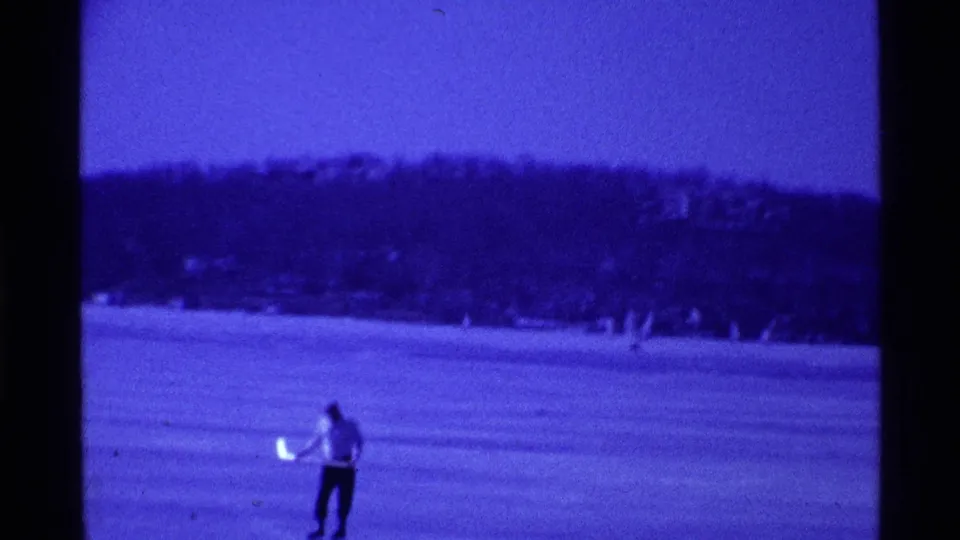 1969 Ohio home-movie frame showing a person skating across a blue-tinted frozen lake with wooded hills behind.