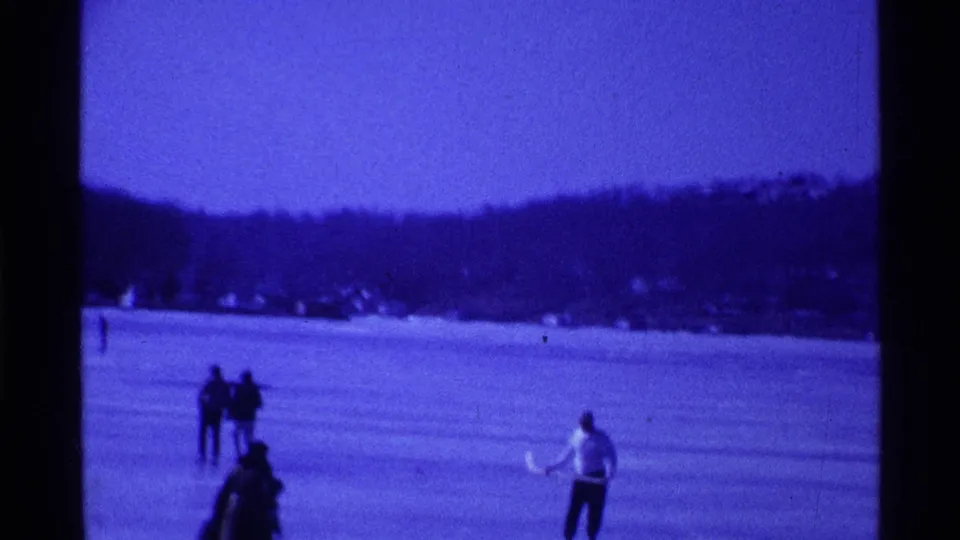 Blue-tinted 1969 Ohio 8mm frame with scattered people on a frozen lake and dark wooded hills beyond.