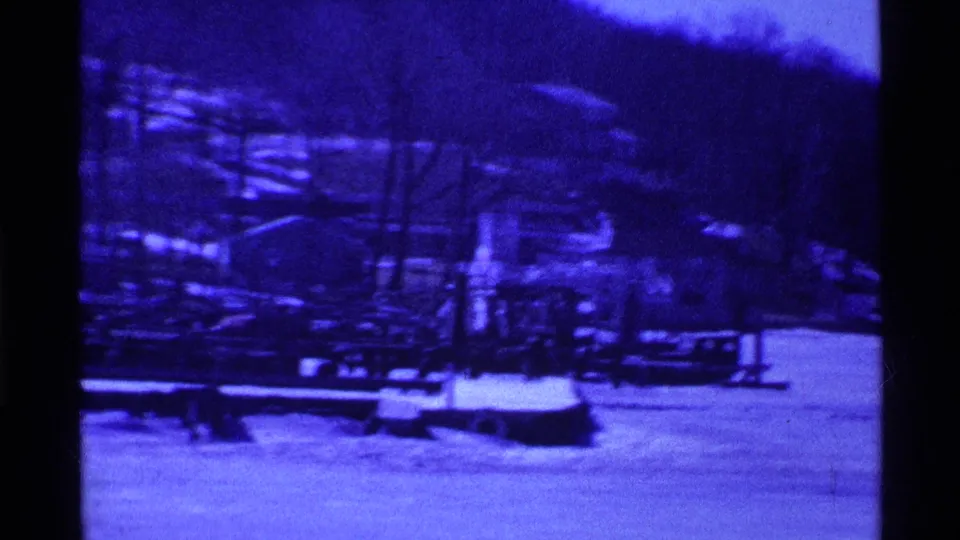 Blue-shifted 1969 Ohio home-movie frame of a snowy park or lakeside area with railings, bare trees, and distant hillside.