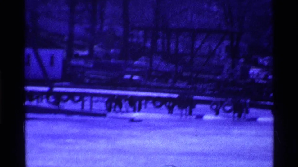 Blue-tinted 1969 Ohio home-movie frame showing snowy ground, dark benches or railing forms, bare trees, and park structures.